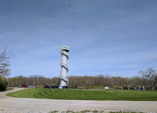 A twisting observation tower stands in a park, with visitors and cars nearby under a clear blue sky.