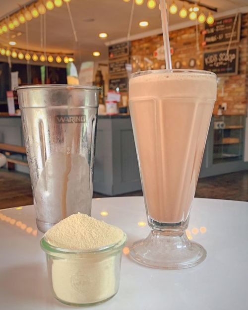 A milkshake in a tall glass next to a shaker and a small jar of powder, set in a cozy café with warm lighting.