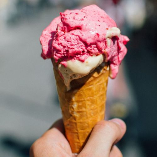 A hand holding a cone with pink strawberry ice cream on top, melting slightly in the sunlight.