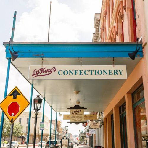 Sign for La King's Confectionery hanging above a storefront, with a street sign and lamp post visible below.