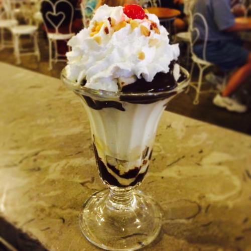 A tall glass dessert featuring layers of ice cream, chocolate sauce, whipped cream, nuts, and a cherry on top.