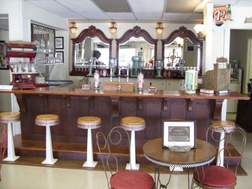 A vintage soda fountain bar with wooden counter, stools, and retro decor, featuring mirrors and nostalgic drink dispensers.