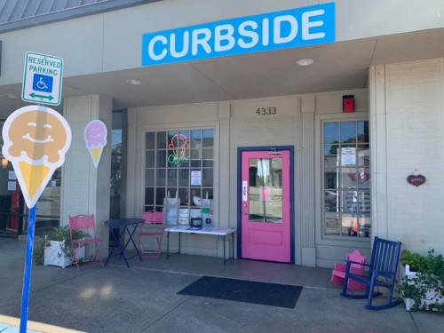 A colorful storefront with a sign reading "CURBSIDE," featuring pink doors and outdoor seating with ice cream signs.