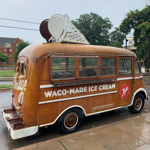 A vintage ice cream truck with a large ice cream cone on top, parked on a rainy street, promoting Waco-made ice cream.