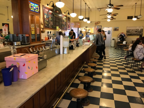 A vintage diner interior with a long marble counter, stools, and checkered floor, bustling with customers and staff.
