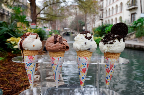 Four ice cream cones in a row, featuring chocolate, vanilla, and cookies & cream flavors, set by a scenic canal.