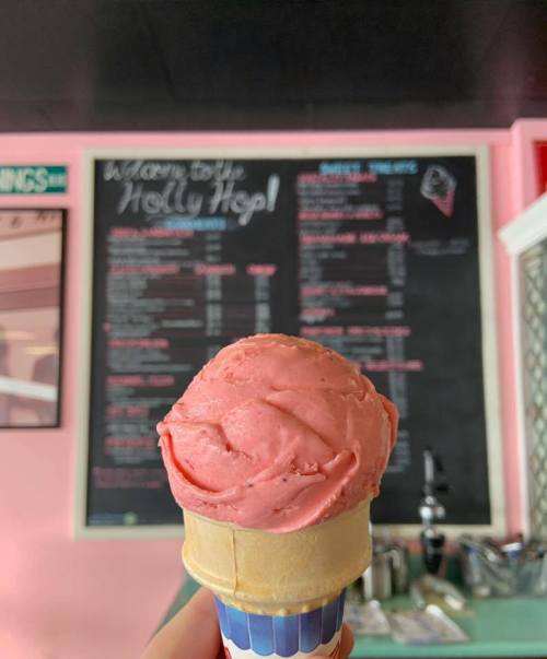 A hand holds a cone of pink ice cream in front of a chalkboard menu at an ice cream shop.