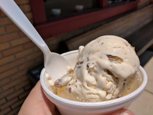 A hand holding a cup of creamy ice cream with chunks and a plastic spoon resting inside.