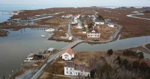 Aerial view of a coastal village with houses, marshland, and winding waterways. Boats are visible in the water.