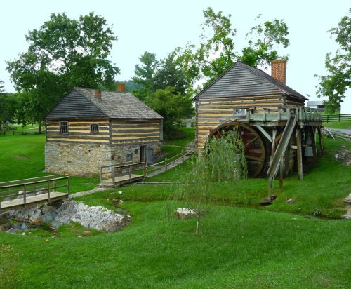 Historic wooden buildings with a water wheel, surrounded by green grass and trees, near a small stream.