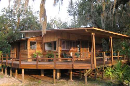 A wooden cabin on stilts with a spacious porch, surrounded by trees and greenery.