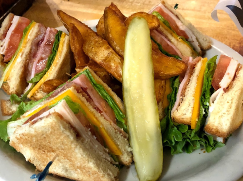 A plate with a club sandwich, crispy potato wedges, and a pickle slice.