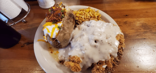 A plate featuring fried chicken smothered in gravy, a loaded baked potato, and a side of corn.
