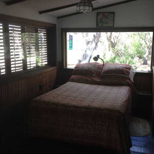 Cozy bedroom with a patterned bedspread, wooden walls, and a large window overlooking greenery.