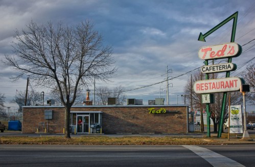 A brick building with a sign reading "Ted's Cafeteria Restaurant" under a cloudy sky, featuring a retro arrow design.