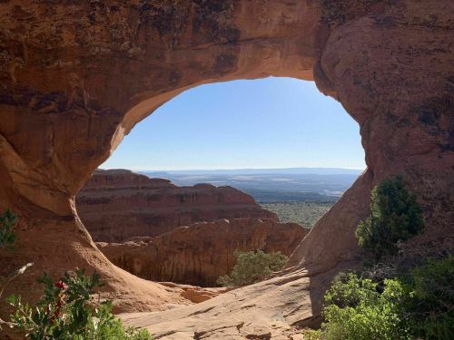 A natural rock arch frames a scenic view of layered red rock formations and distant mountains under a clear blue sky.