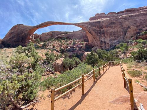Natural rock arch towering over a sandy path, surrounded by greenery and rugged terrain under a blue sky.