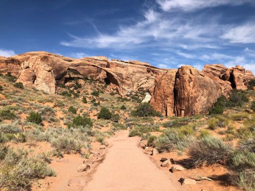 A sandy path leads through a desert landscape with large rock formations and a blue sky with wispy clouds.
