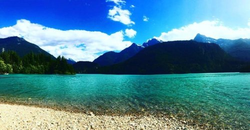 A serene lake surrounded by mountains, with clear turquoise water and a bright blue sky dotted with clouds.