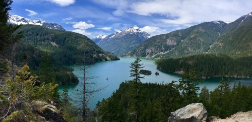 A panoramic view of a turquoise lake surrounded by lush green forests and snow-capped mountains under a blue sky.