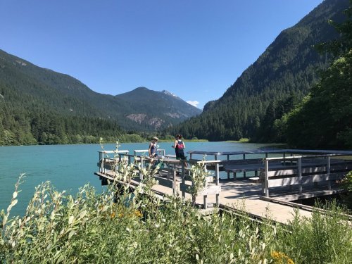 Two people stand on a wooden dock by a turquoise lake, surrounded by lush green mountains under a clear blue sky.