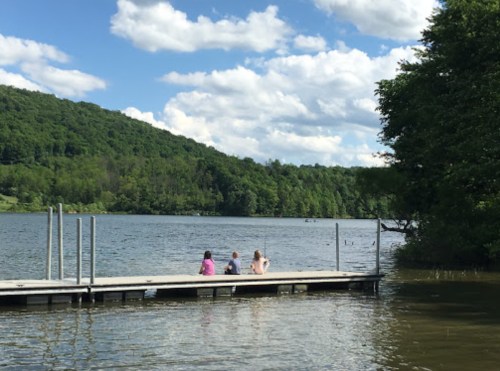 Three people sit on a dock by a lake, surrounded by green hills and a partly cloudy sky.