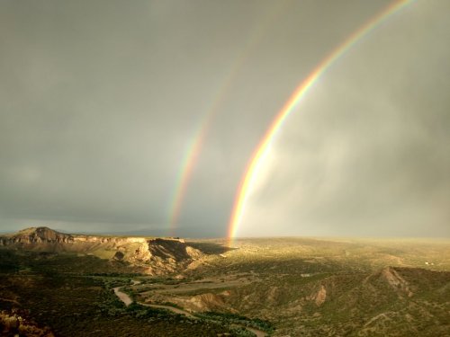 A scenic view of a double rainbow arching over a green valley and winding river under a cloudy sky.