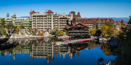 A scenic view of a large, historic hotel by a lake, surrounded by trees and mountains under a clear blue sky.