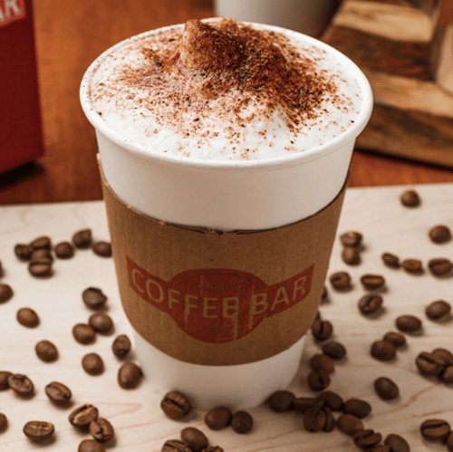 A coffee cup topped with frothy cream and sprinkled with cocoa, surrounded by coffee beans on a wooden surface.