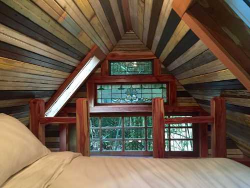 Cozy attic interior with wooden walls, stained glass window, and a view of greenery outside.