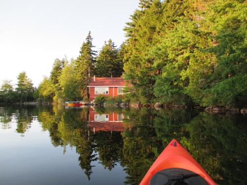 A red cabin by a calm lake, surrounded by lush green trees, with reflections on the water and a kayak in the foreground.
