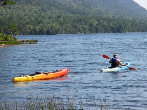 A person in a blue kayak paddles alongside a tethered orange kayak on a calm lake surrounded by trees.