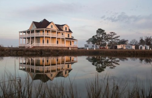 A yellow house with a porch reflects in calm water, surrounded by trees and a clear sky at dusk.