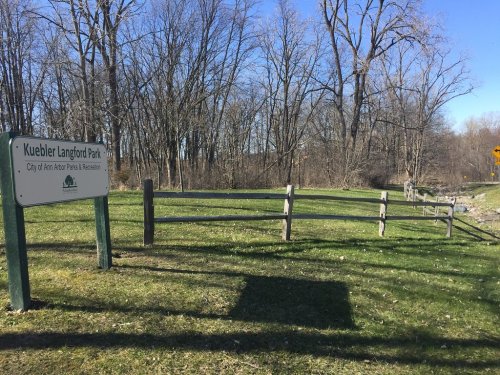 Sign for Kuebler Langford Park, surrounded by trees and grassy area, with a winding path nearby.