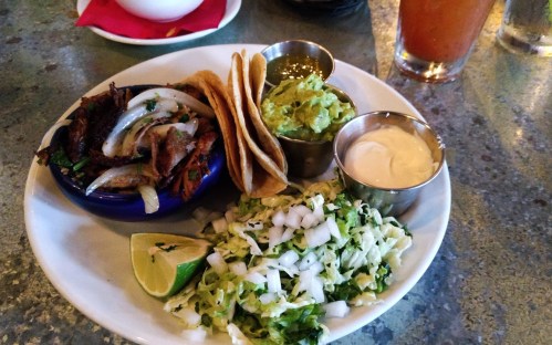A plate of tacos with grilled meat, onions, guacamole, sour cream, and a side of shredded lettuce and lime.