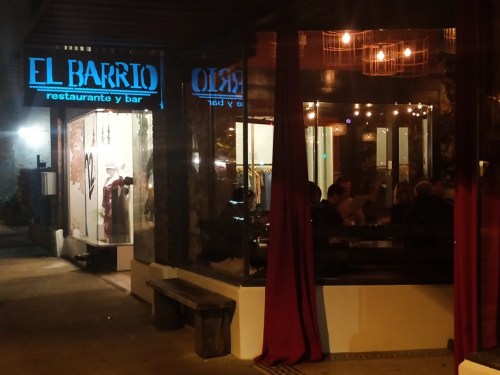 A nighttime view of "El Barrio" restaurant and bar, featuring warm lights and a cozy outdoor seating area.