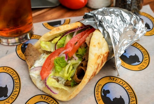A wrapped sandwich filled with lettuce, tomato, onion, and meat, served on a branded paper with a drink in the background.