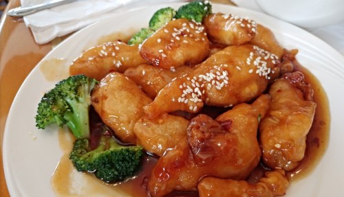 A plate of sesame chicken with broccoli, drizzled in a sweet sauce, garnished with sesame seeds.