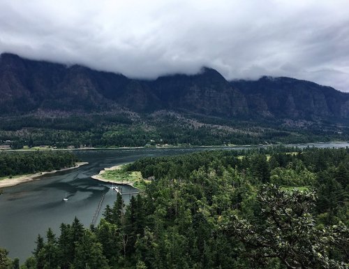 A scenic view of a river winding through lush green forests, with mountains shrouded in clouds in the background.