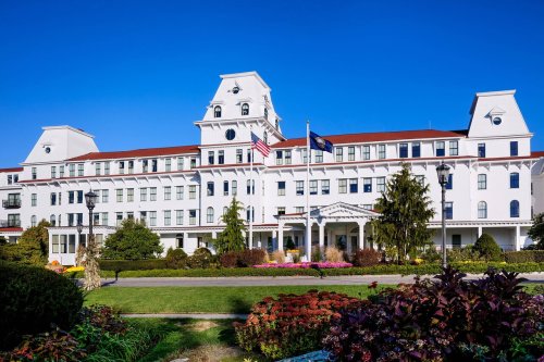 A large white hotel with a red roof, surrounded by colorful gardens and blue sky. Flags are displayed in front.