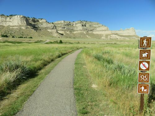 A paved path leads through grassy terrain with rocky cliffs in the background and trail signs on the right.