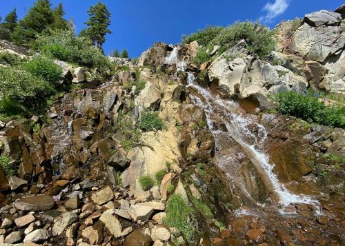 A cascading waterfall flows over rocky terrain, surrounded by lush greenery and a clear blue sky.
