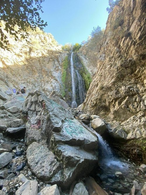 A waterfall cascades down rocky cliffs, surrounded by greenery and boulders, with people in the distance enjoying the view.