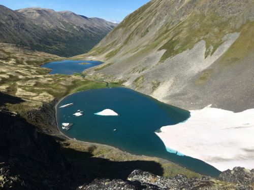 Aerial view of two glacial lakes surrounded by mountains and rocky terrain, with patches of snow and ice visible.