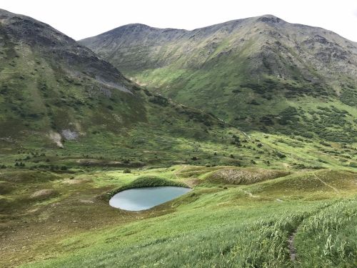 A serene mountain landscape featuring a small, circular pond surrounded by lush green hills and peaks in the background.