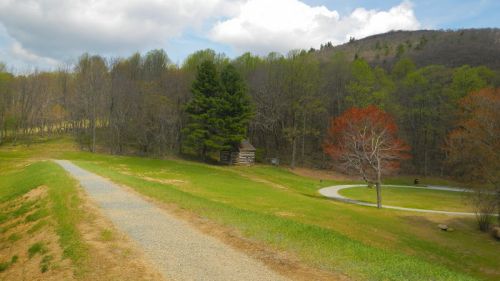 A winding path leads through a green landscape with trees and a small cabin in the distance under a cloudy sky.