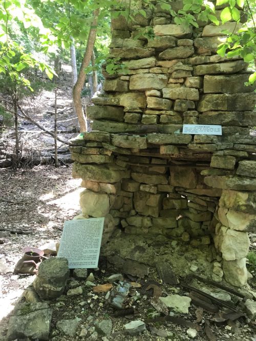 Stone chimney ruins surrounded by trees, with a plaque and scattered debris on the ground.