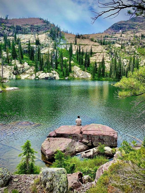 A person sits on a rock by a serene lake, surrounded by lush greenery and mountains in the background.