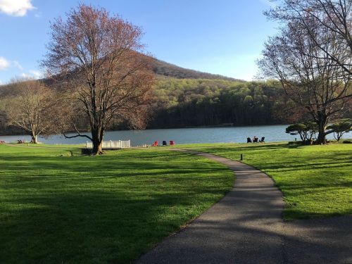 A serene lake surrounded by green hills, with a winding path and trees, under a clear blue sky.
