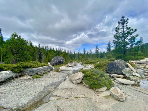 A rocky landscape with a stream, surrounded by lush green trees and a cloudy sky.
